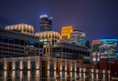 A night view of downtown Minneapolis glows with vibrant hues from its illuminated skyline. The IDS Center and Wells Fargo Tower stand prominently among the city’s modern high-rises, their lights reflecting the urban energy below. New residential buildings in the foreground contrast with the historic clock tower in the distance, highlighting the city’s mix of old and new architecture.