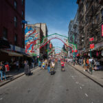 People walk along Mulberry Street in Manhattan’s Little Italy under rows of simple festive arches. Shops and cafés line the street as vendors sell food and souvenirs to passersby. The scene reflects the neighborhood’s everyday rhythm and long-standing cultural roots.
