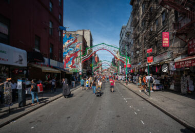 People walk along Mulberry Street in Manhattan’s Little Italy under rows of simple festive arches. Shops and cafés line the street as vendors sell food and souvenirs to passersby. The scene reflects the neighborhood’s everyday rhythm and long-standing cultural roots.