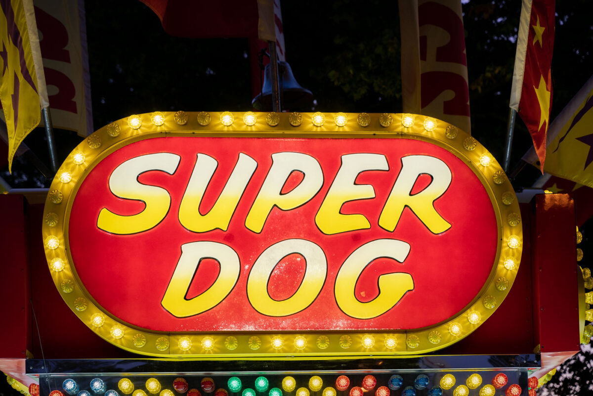 A brightly lit “Super Dog” food stand sign glows with yellow bulbs at a carnival or fairground. Surrounded by flags and festive colors, the concession stand evokes the classic atmosphere of American fairs where hot dogs and snacks are a staple of the midway.