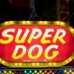 A brightly lit “Super Dog” food stand sign glows with yellow bulbs at a carnival or fairground. Surrounded by flags and festive colors, the concession stand evokes the classic atmosphere of American fairs where hot dogs and snacks are a staple of the midway.