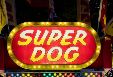 A brightly lit “Super Dog” food stand sign glows with yellow bulbs at a carnival or fairground. Surrounded by flags and festive colors, the concession stand evokes the classic atmosphere of American fairs where hot dogs and snacks are a staple of the midway.