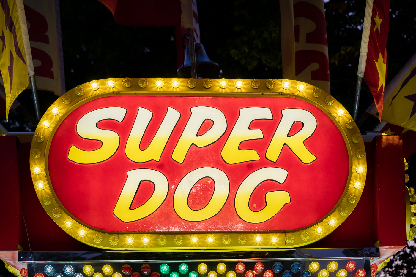 A brightly lit “Super Dog” food stand sign glows with yellow bulbs at a carnival or fairground. Surrounded by flags and festive colors, the concession stand evokes the classic atmosphere of American fairs where hot dogs and snacks are a staple of the midway.
