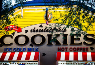 A brightly painted cookie shop features bold signage advertising chocolate chip cookies, ice cold milk, and hot coffee. The storefront, with its yellow siding, red-and-white awnings, and whimsical design, captures the festive atmosphere typical of fairgrounds and boardwalk-style concessions.