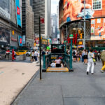 Pedestrians crowd the sidewalks around a subway entrance in Times Square, Manhattan. The 42nd Street–Times Square station, one of the busiest in New York City, connects multiple subway lines beneath the bright billboards, storefronts, and heavy traffic of the theater district.
