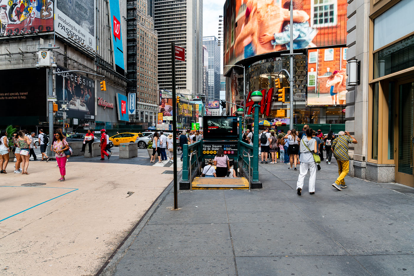 Pedestrians crowd the sidewalks around a subway entrance in Times Square, Manhattan. The 42nd Street–Times Square station, one of the busiest in New York City, connects multiple subway lines beneath the bright billboards, storefronts, and heavy traffic of the theater district.