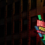 A neon sign for Angelo’s Pizza glows against the brick facade of a building in Midtown Manhattan. The Italian restaurant, located near the Ed Sullivan Theater, adds to the neighborhood’s mix of nightlife and dining spots illuminated after dark.