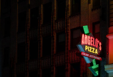 A neon sign for Angelo’s Pizza glows against the brick facade of a building in Midtown Manhattan. The Italian restaurant, located near the Ed Sullivan Theater, adds to the neighborhood’s mix of nightlife and dining spots illuminated after dark.