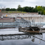 Wastewater treatment plant with sedimentation tanks used to settle and remove solids from incoming water. Metal bridges and railings provide access for workers to maintain and inspect equipment. In the background, storage areas and treatment structures are surrounded by trees and fencing at the edge of the facility.