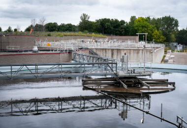 Wastewater treatment plant with sedimentation tanks used to settle and remove solids from incoming water. Metal bridges and railings provide access for workers to maintain and inspect equipment. In the background, storage areas and treatment structures are surrounded by trees and fencing at the edge of the facility.