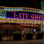 The marquee of the Ed Sullivan Theater glows at night in Manhattan, advertising “The Late Show with Stephen Colbert.” Pedestrians walk past the historic theater, which has been home to late-night television for decades, with nearby businesses like Angelo’s Pizza adding to the busy Midtown street scene.