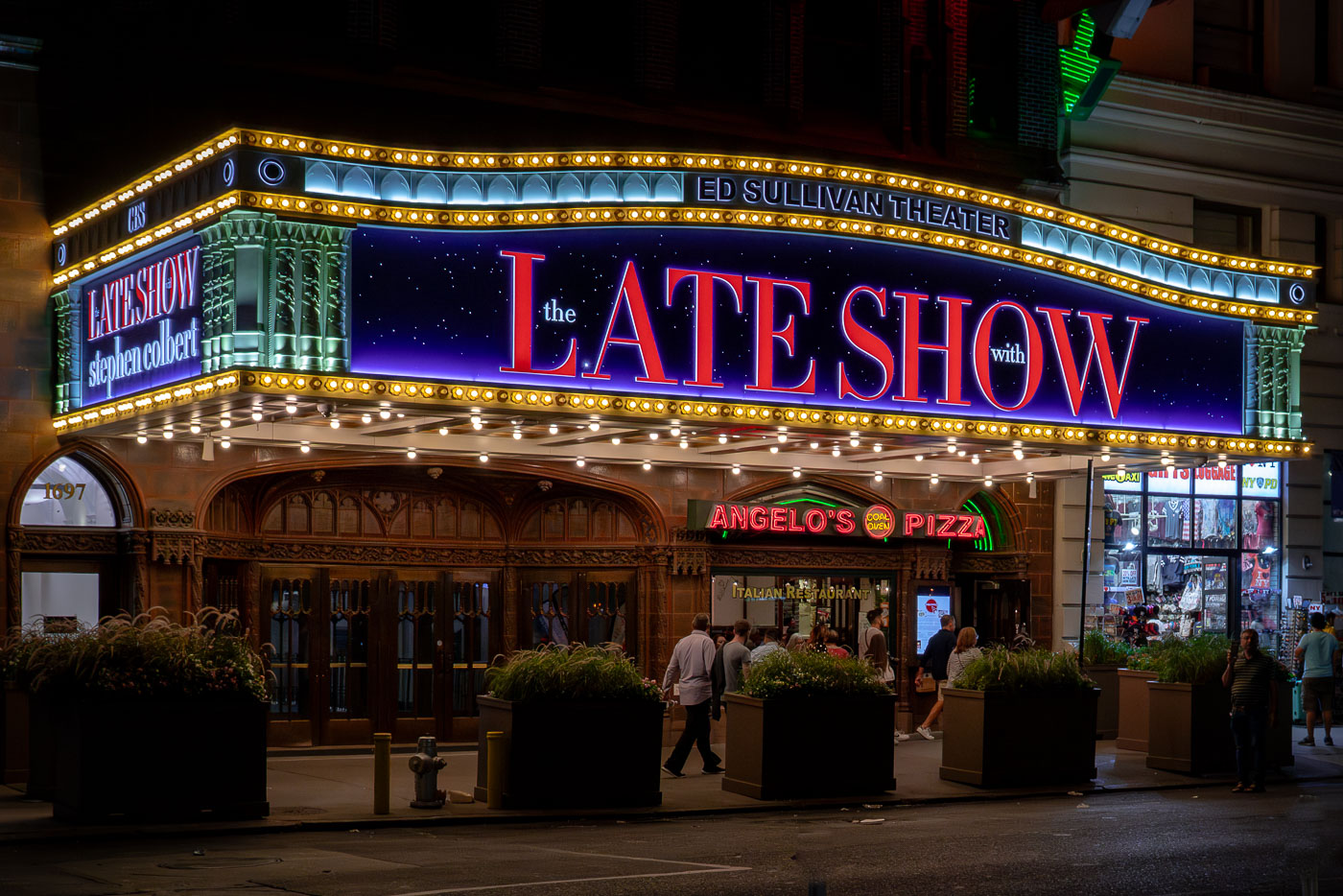 The marquee of the Ed Sullivan Theater glows at night in Manhattan, advertising “The Late Show with Stephen Colbert.” Pedestrians walk past the historic theater, which has been home to late-night television for decades, with nearby businesses like Angelo’s Pizza adding to the busy Midtown street scene.