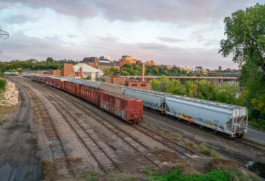 Freight cars sit on rail tracks near the Mississippi River in Minneapolis, with the University of Minnesota campus in the background. The Southeast Steam Plant and its red exhaust stacks rise behind the trains, and the Washington Avenue Bridge crosses the river farther upstream. The scene highlights the mix of industry, infrastructure, and campus development along the riverfront.