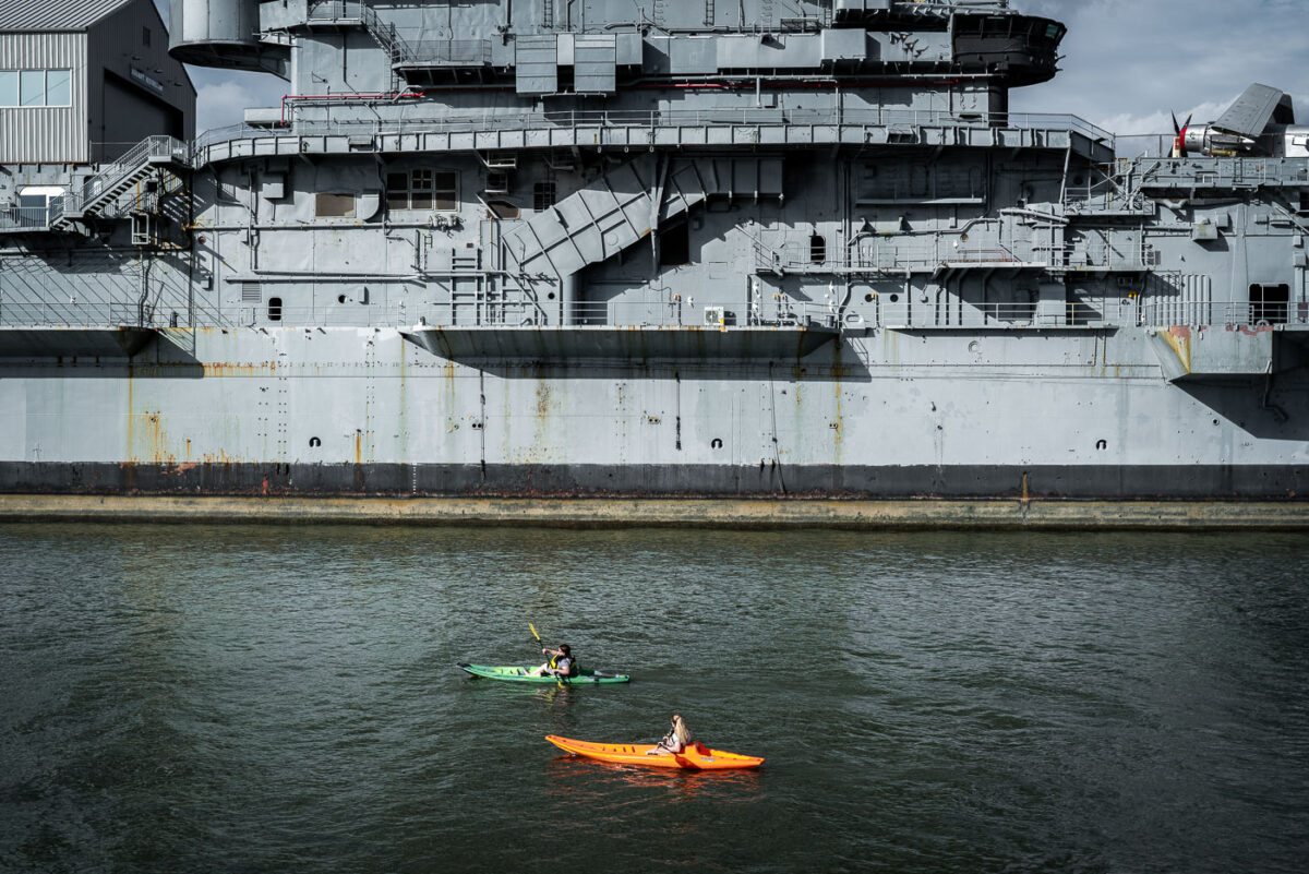 Two people paddle kayaks along the Hudson River near the USS Intrepid, a decommissioned aircraft carrier now serving as part of the Intrepid Sea, Air & Space Museum in Manhattan. The massive gray hull of the ship looms over the water, contrasting with the small boats below.