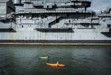 Two people paddle kayaks along the Hudson River near the USS Intrepid, a decommissioned aircraft carrier now serving as part of the Intrepid Sea, Air & Space Museum in Manhattan. The massive gray hull of the ship looms over the water, contrasting with the small boats below.