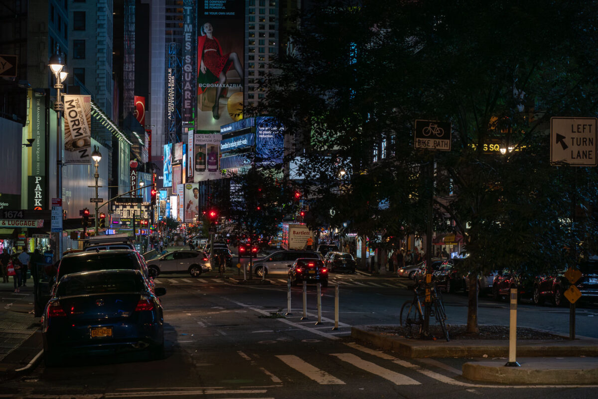 Traffic and illuminated billboards fill Times Square in Manhattan at night, with streams of cars and taxis moving through the crowded intersection. Bright advertisements, theater signs, and neon lights dominate the scene as pedestrians navigate the sidewalks and bike lanes around one of New York City’s busiest landmarks.