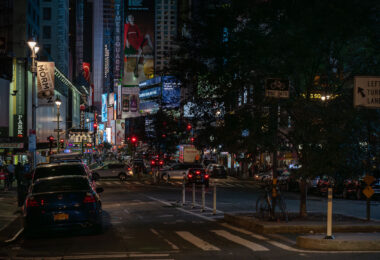 Traffic and illuminated billboards fill Times Square in Manhattan at night, with streams of cars and taxis moving through the crowded intersection. Bright advertisements, theater signs, and neon lights dominate the scene as pedestrians navigate the sidewalks and bike lanes around one of New York City’s busiest landmarks.