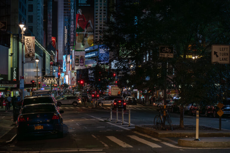 Manhattan advertising at night 3 Traffic and illuminated billboards fill Times Square in Manhattan at night, with streams of cars and taxis moving through the crowded intersection. Bright advertisements, theater signs, and neon lights dominate the scene as pedestrians navigate the sidewalks and bike lanes around one of New York City’s busiest landmarks.