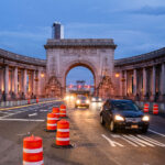 Cars pass through the Manhattan Bridge arch and colonnade at dusk in Lower Manhattan on Sunday, Sept. 14, 2025. Orange traffic barrels line the roadway as construction continues near the historic Beaux-Arts structure.