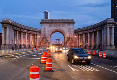 Cars pass through the Manhattan Bridge arch and colonnade at dusk in Lower Manhattan on Sunday, Sept. 14, 2025. Orange traffic barrels line the roadway as construction continues near the historic Beaux-Arts structure.