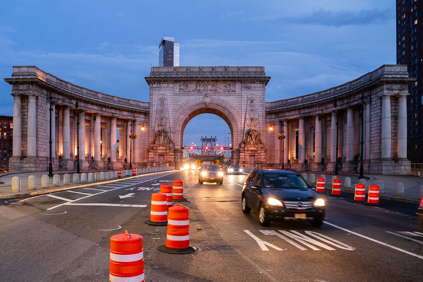 Cars pass through the Manhattan Bridge arch and colonnade at dusk in Lower Manhattan on Sunday, Sept. 14, 2025. Orange traffic barrels line the roadway as construction continues near the historic Beaux-Arts structure.
