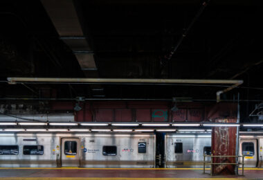 A Metro-North Railroad train sits at a platform inside Grand Central Terminal in Manhattan. The stainless-steel cars reflect the dim lighting of the underground station, where exposed pipes and graffiti-marked beams highlight the aging infrastructure of the busy commuter hub.