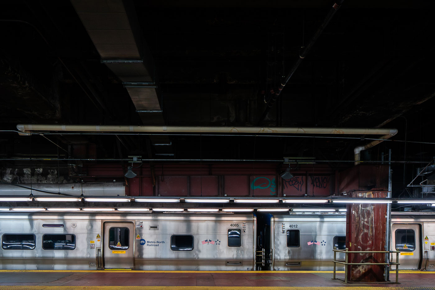 A Metro-North Railroad train sits at a platform inside Grand Central Terminal in Manhattan. The stainless-steel cars reflect the dim lighting of the underground station, where exposed pipes and graffiti-marked beams highlight the aging infrastructure of the busy commuter hub.