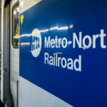 A Metro-North Railroad train is seen at a station platform, its blue-and-white exterior bearing the MTA logo. The commuter rail line, operated by the Metropolitan Transportation Authority, connects New York City with suburbs across New York State and Connecticut.