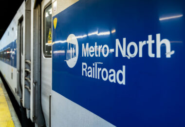 A Metro-North Railroad train is seen at a station platform, its blue-and-white exterior bearing the MTA logo. The commuter rail line, operated by the Metropolitan Transportation Authority, connects New York City with suburbs across New York State and Connecticut.