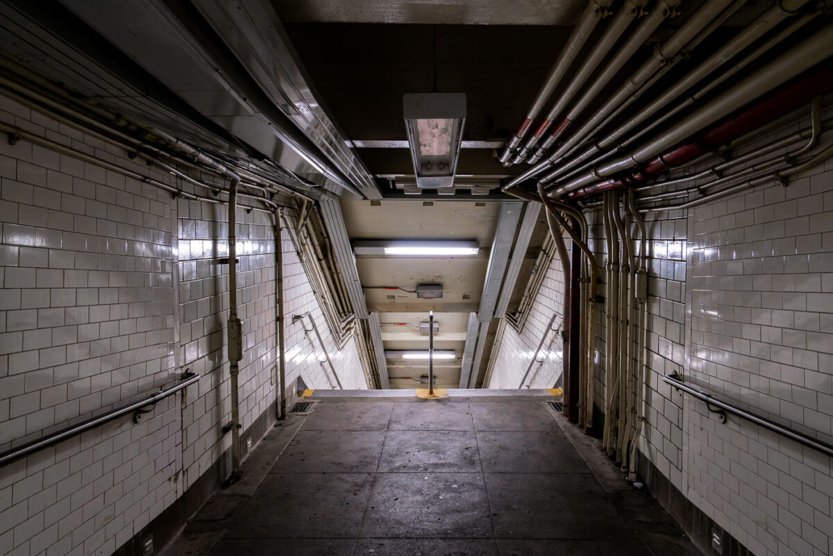 A dimly lit stairway leads down into a New York City subway station on Sunday, Sept. 14, 2025. Aging infrastructure and exposed piping are visible along the tiled walls.