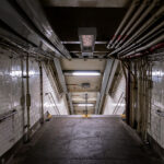 A dimly lit stairway leads down into a New York City subway station on Sunday, Sept. 14, 2025. Aging infrastructure and exposed piping are visible along the tiled walls.