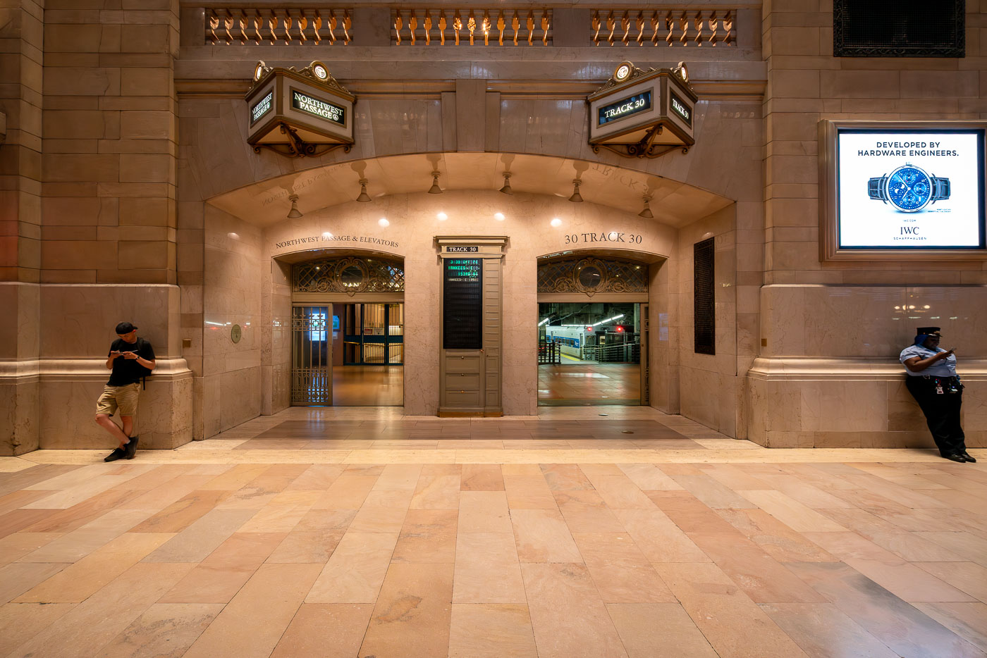 The entrance to Track 30 inside Grand Central Terminal in Manhattan is seen at night. The marble hallways of the landmark station lead to the train platforms below, as a commuter and a security officer stand near the archways beneath illuminated signs.