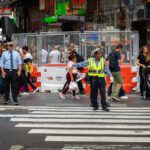 A traffic officer directs pedestrians at a crosswalk in Times Square, Manhattan. Crowds move past construction barriers and street signage in the busy tourist district, where theaters, billboards, and entertainment venues draw thousands of visitors daily.