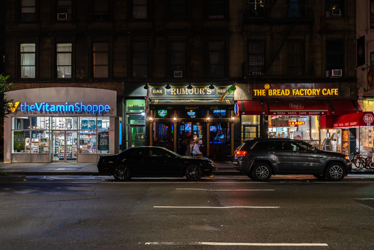 Storefronts of The Vitamin Shoppe, Rumour’s Bar and Grill, and The Bread Factory Café are seen at night along a Manhattan street. Cars are parked in front of the businesses.