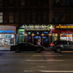 Storefronts of The Vitamin Shoppe, Rumour’s Bar and Grill, and The Bread Factory Café are seen at night along a Manhattan street. Cars are parked in front of the businesses.