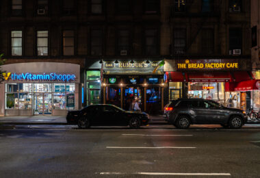 Storefronts of The Vitamin Shoppe, Rumour’s Bar and Grill, and The Bread Factory Café are seen at night along a Manhattan street. Cars are parked in front of the businesses.