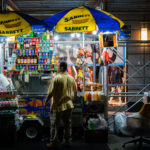 A vendor works at a brightly lit Sabrett hot dog stand in Manhattan, surrounded by stacks of soda cans, bottled water, and snack foods. Street carts like these are a common sight across New York City, serving pretzels, knishes, and hot dogs late into the night to passing pedestrians.
