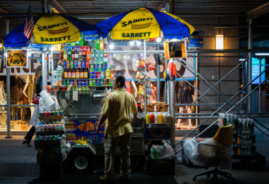 A vendor works at a brightly lit Sabrett hot dog stand in Manhattan, surrounded by stacks of soda cans, bottled water, and snack foods. Street carts like these are a common sight across New York City, serving pretzels, knishes, and hot dogs late into the night to passing pedestrians.