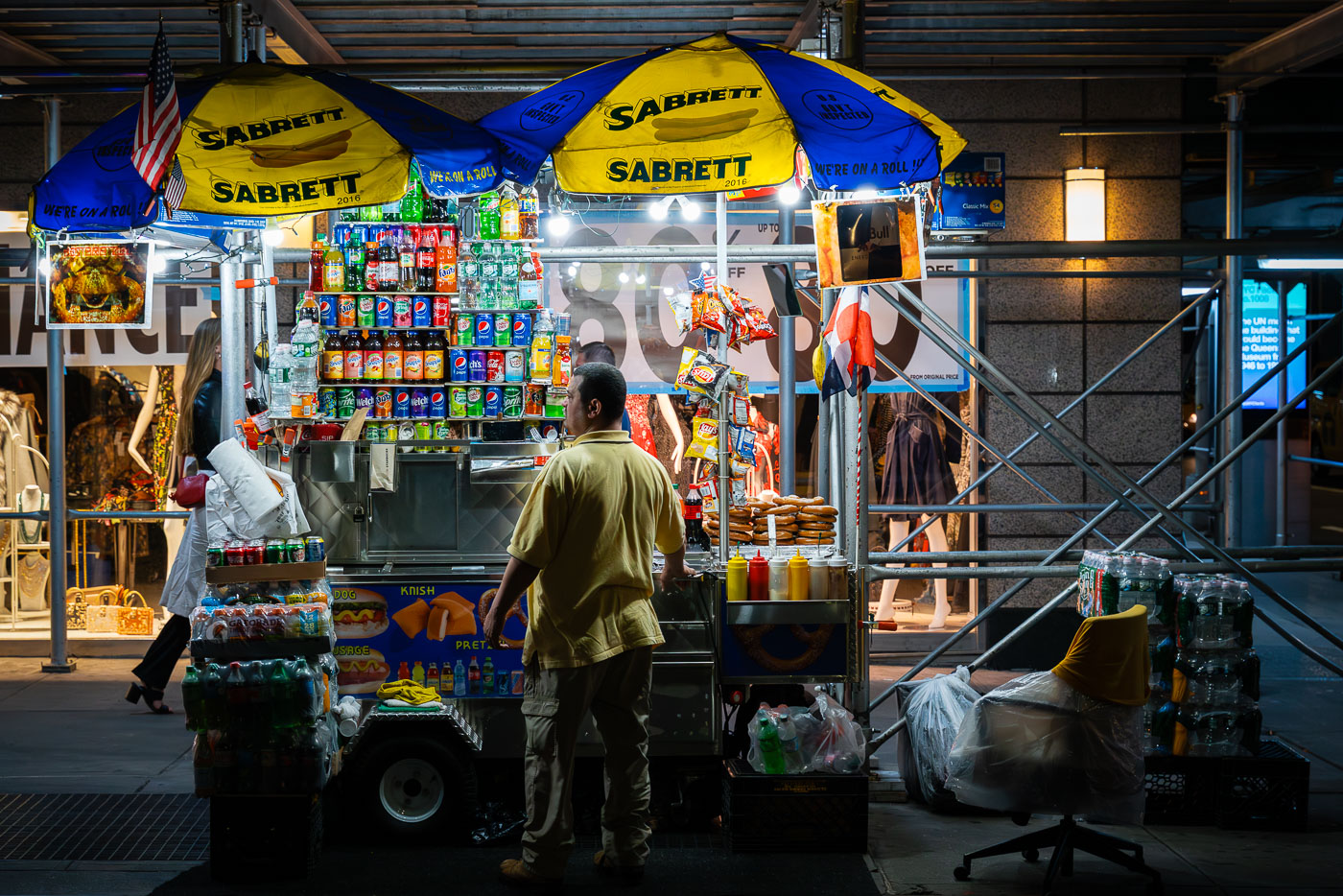 A vendor works at a brightly lit Sabrett hot dog stand in Manhattan, surrounded by stacks of soda cans, bottled water, and snack foods. Street carts like these are a common sight across New York City, serving pretzels, knishes, and hot dogs late into the night to passing pedestrians.
