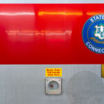 A Connecticut state seal is displayed on the side of a Metro-North Railroad train car, operated in partnership with the Connecticut Department of Transportation. The red-and-silver exterior includes safety instructions and signage, highlighting the regional rail system that connects commuters between New York and Connecticut.