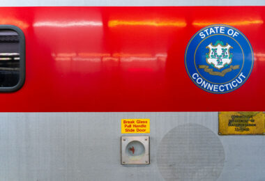 A Connecticut state seal is displayed on the side of a Metro-North Railroad train car, operated in partnership with the Connecticut Department of Transportation. The red-and-silver exterior includes safety instructions and signage, highlighting the regional rail system that connects commuters between New York and Connecticut.