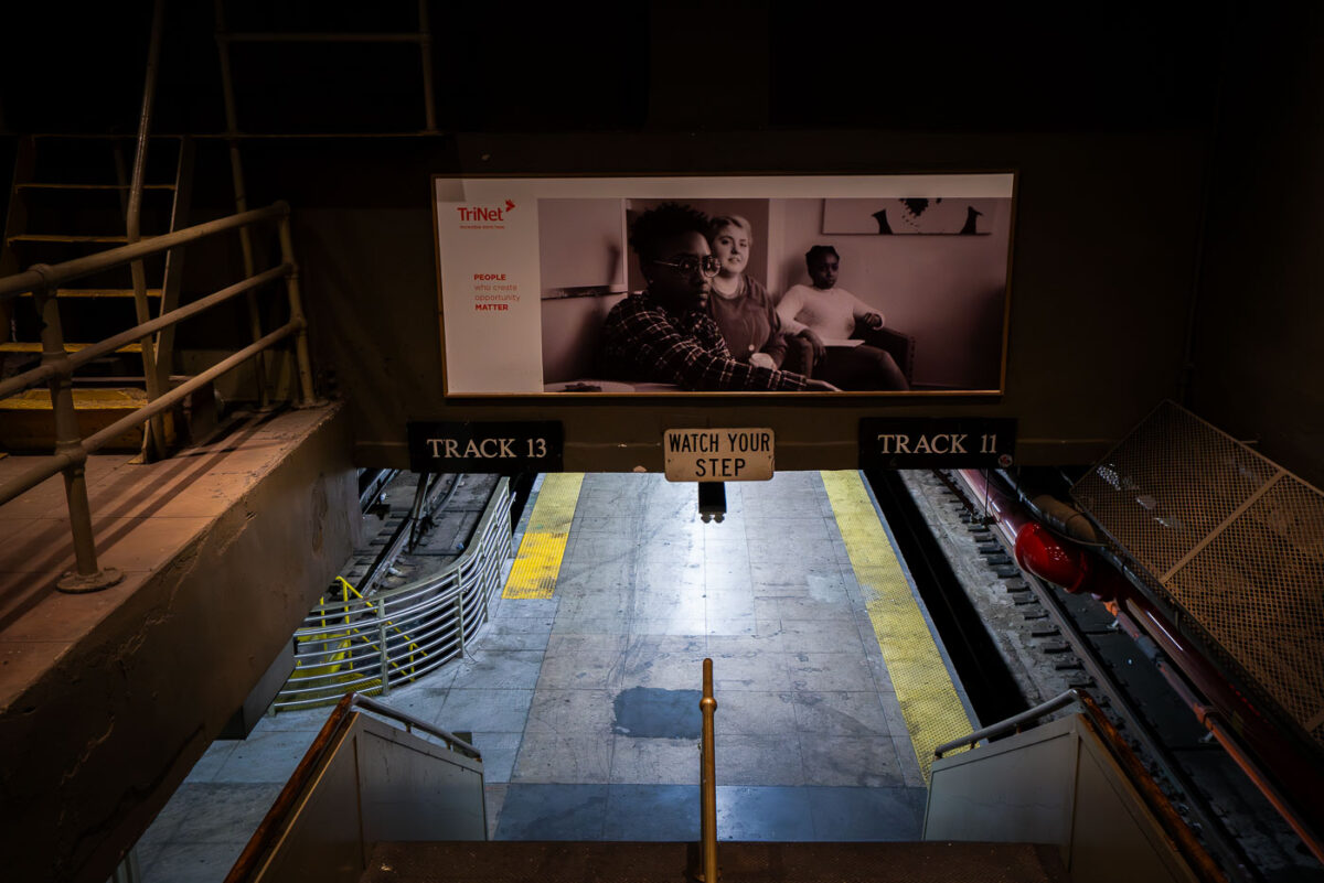 A stairway leads down to the train platforms for Tracks 11 and 13 at Penn Station in Manhattan. A sign reading “Watch Your Step” hangs above the stairs, while a black-and-white advertisement looms over the tracks in the busy transit hub.