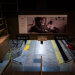 A stairway leads down to the train platforms for Tracks 11 and 13 at Penn Station in Manhattan. A sign reading “Watch Your Step” hangs above the stairs, while a black-and-white advertisement looms over the tracks in the busy transit hub.