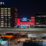 Opened in 2014, Target Field Station is a multimodal transit hub serving the METRO Blue and Green light rail lines, Northstar commuter rail, and several Metro Transit bus routes. It sits adjacent to Target Field, home of the Minnesota Twins, and doubles as a public gathering space with an amphitheater, green plaza, and year-round programming.In this night view, the glowing red canopy and bold “Target Field Station” signage stand out against the dark sky, framed between the historic Ford Center (left, built in 1912 as an assembly plant) and a modern office building (right). A freeway bridge cuts through the foreground, symbolizing the layered infrastructure of the city.This mix of historic brickwork, modern transit, and illuminated signage captures Minneapolis’s ongoing balance of industrial heritage and urban redevelopment.