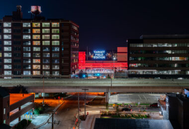 Opened in 2014, Target Field Station is a multimodal transit hub serving the METRO Blue and Green light rail lines, Northstar commuter rail, and several Metro Transit bus routes. It sits adjacent to Target Field, home of the Minnesota Twins, and doubles as a public gathering space with an amphitheater, green plaza, and year-round programming.

In this night view, the glowing red canopy and bold “Target Field Station” signage stand out against the dark sky, framed between the historic Ford Center (left, built in 1912 as an assembly plant) and a modern office building (right). A freeway bridge cuts through the foreground, symbolizing the layered infrastructure of the city.

This mix of historic brickwork, modern transit, and illuminated signage captures Minneapolis’s ongoing balance of industrial heritage and urban redevelopment.