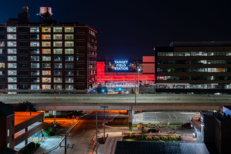 Target Field Station in Minneapolis North Loop 3 Opened in 2014, Target Field Station is a multimodal transit hub serving the METRO Blue and Green light rail lines, Northstar commuter rail, and several Metro Transit bus routes. It sits adjacent to Target Field, home of the Minnesota Twins, and doubles as a public gathering space with an amphitheater, green plaza, and year-round programming.In this night view, the glowing red canopy and bold “Target Field Station” signage stand out against the dark sky, framed between the historic Ford Center (left, built in 1912 as an assembly plant) and a modern office building (right). A freeway bridge cuts through the foreground, symbolizing the layered infrastructure of the city.This mix of historic brickwork, modern transit, and illuminated signage captures Minneapolis’s ongoing balance of industrial heritage and urban redevelopment.