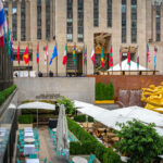 The Lower Plaza at Rockefeller Center in Midtown Manhattan is seen with the golden statue of Prometheus overlooking the outdoor dining area. Rows of international flags line the upper concourse, while crowds gather around the landmark complex that is home to shops, offices, and seasonal attractions.