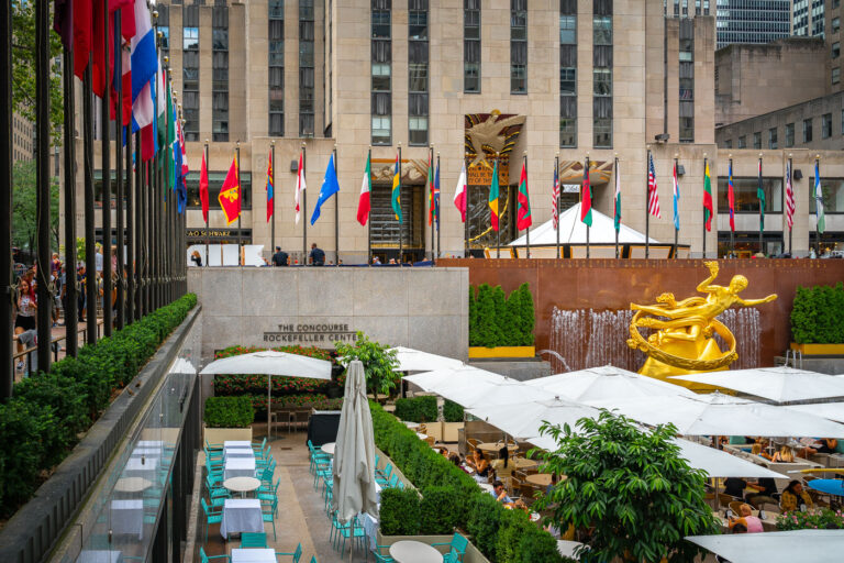 The Concourse at Rockefeller Center 2 The Lower Plaza at Rockefeller Center in Midtown Manhattan is seen with the golden statue of Prometheus overlooking the outdoor dining area. Rows of international flags line the upper concourse, while crowds gather around the landmark complex that is home to shops, offices, and seasonal attractions.