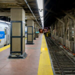 A Metro-North Railroad train waits at a platform inside Grand Central Terminal in Manhattan. The concrete pillars and exposed beams of the underground station frame the tracks, where the yellow warning strip lines the edge of the platform.