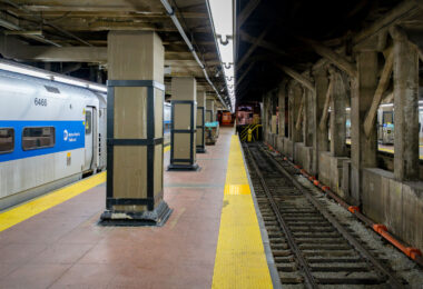 A Metro-North Railroad train waits at a platform inside Grand Central Terminal in Manhattan. The concrete pillars and exposed beams of the underground station frame the tracks, where the yellow warning strip lines the edge of the platform.